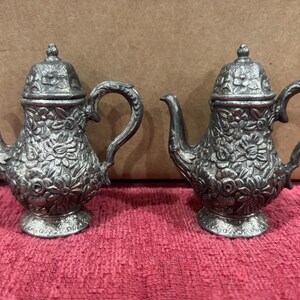 May include: Two antique silver-colored miniature teapots. Each teapot features a floral design, a curved handle, a spout, and a lid with a small knob. The teapots are displayed on a pink surface.