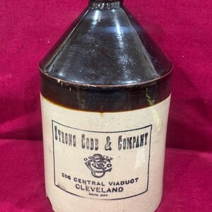May include: Antique stoneware jug with a dark brown glazed top and a cream-colored body. The jug features a rectangular label with the text "STRONG COBB & COMPANY" and the address "206 CENTRAL VIADUCT CLEVELAND". A vintage collectible.