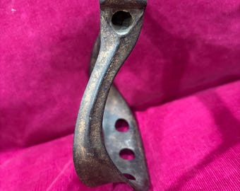 May include: A vintage, bronze-colored stirrup with a curved design. The stirrup has a rectangular top with a circular hole and three circular holes on the side. The metal shows signs of wear, suggesting age and use. The stirrup is likely for equestrian use.