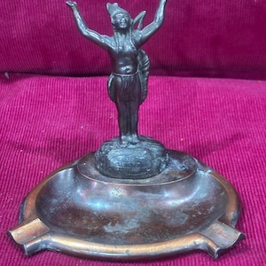 May include: Antique bronze ashtray featuring a Native American figure with arms raised. The ashtray has a circular design with a small platform for the figure and a surrounding dish with cigarette rests. The bronze has a weathered patina.