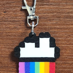 May include: A colorful keychain featuring a pixelated design with a rainbow base and a black and white top. The keychain has a silver clasp for easy attachment. The design is reminiscent of a cupcake or a building.