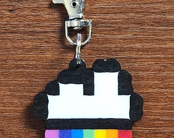 Puede incluir: Un llavero colorido con un diseño pixelado, que presenta una base de arcoíris y una parte superior en blanco y negro. Tiene un cierre plateado para sujetarlo. El diseño recuerda a una magdalena o un edificio.