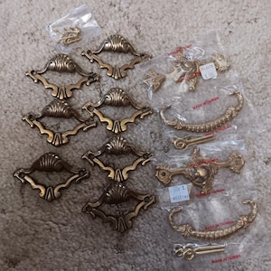 May include: A collection of brass-toned metal drawer pulls and handles. Several pieces feature ornate, shell-like central motifs with decorative arms. Some items are individually packaged in clear plastic bags, with visible screws and labels.
