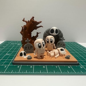 May include: A Halloween-themed diorama featuring cartoon skeleton figures, tombstones, and a spooky tree. The scene includes a gray archway with a skull design, and a sandy base. The figures have large, expressive eyes, and the tombstones read "R.I.P."
