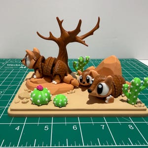 Puede incluir: Una escena desértica caprichosa con tres figuras de armadillo, un árbol desnudo y cactus. Los armadillos son marrones con ojos grandes, colocados sobre una base beige. Los cactus son verdes con detalles blancos y una flor rosa.