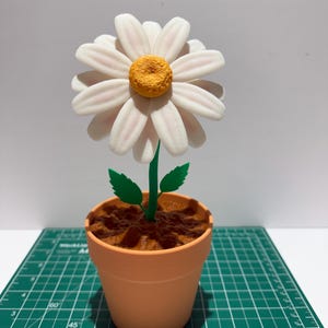 May include: A 3D-printed daisy flower in a terracotta-coloured pot. The flower has white petals with pink accents and a yellow centre. The green stem has two leaves. The pot sits on a green cutting mat.