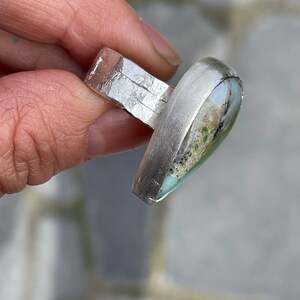 May include: A silver ring with a rectangular band and a large, teardrop-shaped stone. The stone features a mix of colors, including green, white, and black. The ring is held in a hand, showcasing its design and craftsmanship.