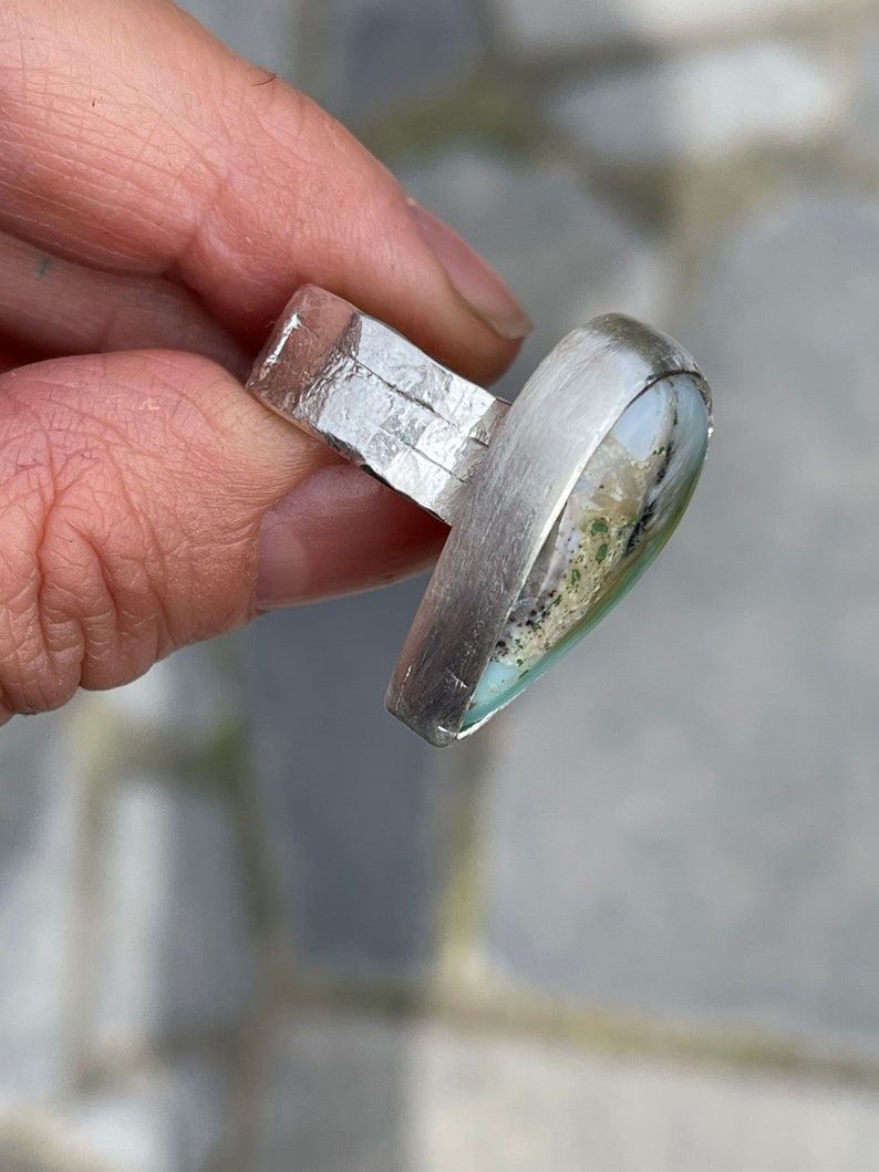 May include: A silver ring with a rectangular band and a large, teardrop-shaped stone. The stone features a mix of colors, including green, white, and black. The ring is held in a hand, showcasing its design and craftsmanship.