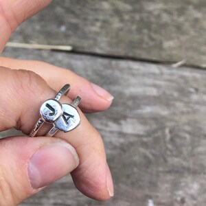 May include: A silver ring featuring two circular pendants, each with a single initial. One pendant displays the letter "J," and the other shows "A." The ring has a textured band and a simple design, suitable for everyday wear.