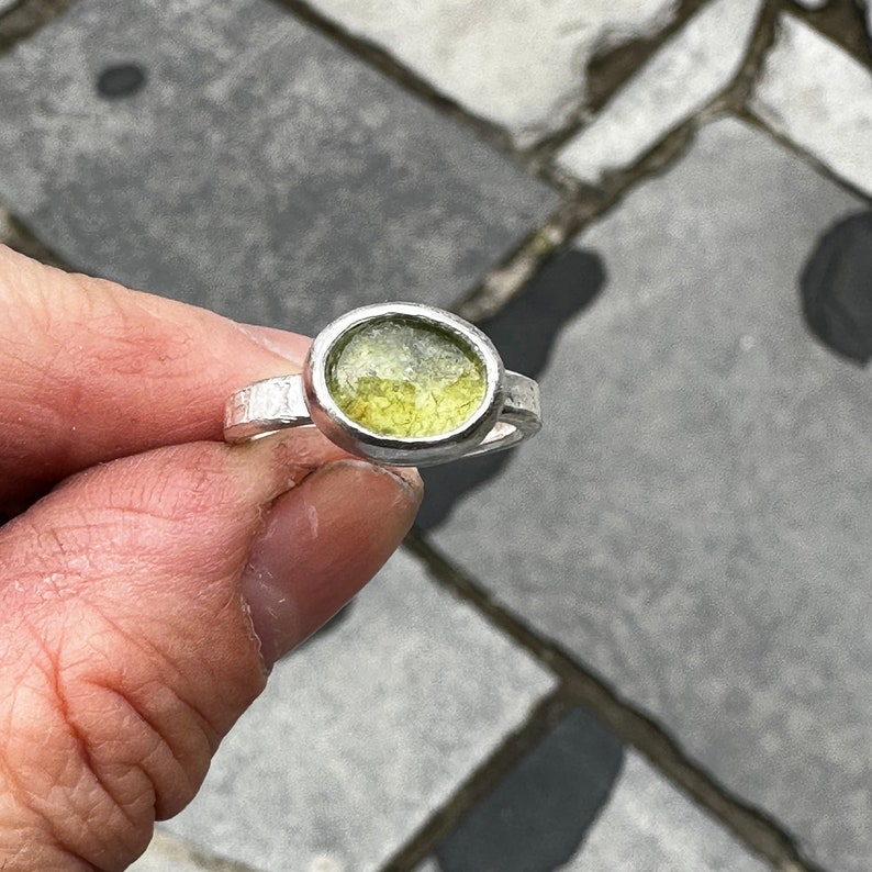 May include: A silver ring featuring a bezel-set, oval-shaped, translucent light green stone. The metal band has a textured, hammered finish, providing a distinct contrast to the smooth, polished silver setting that secures the central gemstone.