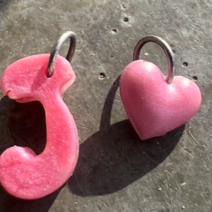 Może przedstawiać: Dwa różowe charmsy z żywicy, jeden w kształcie serca, a drugi w kształcie słuchawki telefonicznej, oba z srebrnymi kółkami.