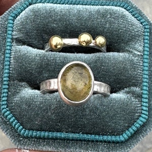 May include: Two silver rings displayed in a teal velvet box. The top ring features three small gold spheres on a hammered silver band. The bottom ring has an oval-shaped, translucent olive-green stone set in a hammered silver band.