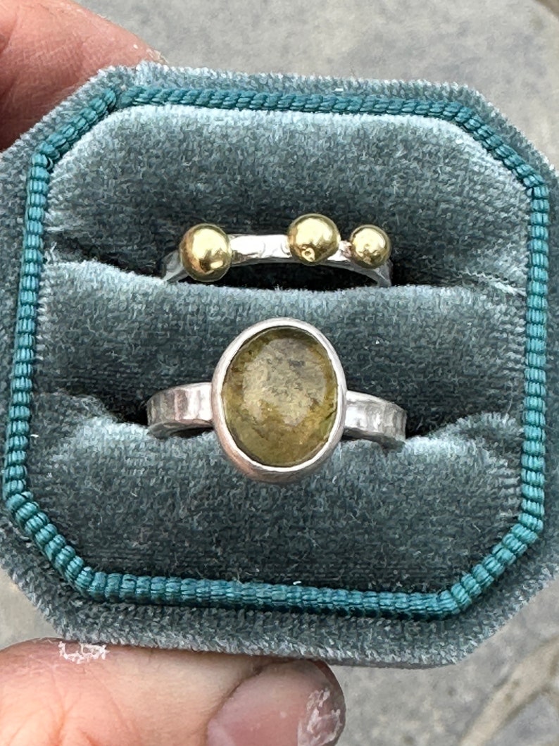 May include: Two silver rings displayed in a teal velvet box. The top ring features three small gold spheres on a hammered silver band. The bottom ring has an oval-shaped, translucent olive-green stone set in a hammered silver band.