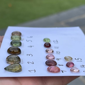 May include: A collection of fifteen loose, polished cabochon gemstones arranged in three vertical columns on a white card. The stones display a variety of colors including green, brown, pink, and clear tones, each labeled with a handwritten number.