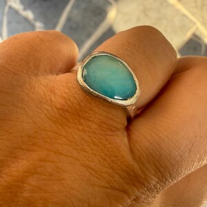 May include: A silver ring with a large, oval-shaped, light blue stone. The stone has a smooth, polished surface and is set in a textured silver band. The ring is displayed on a hand.