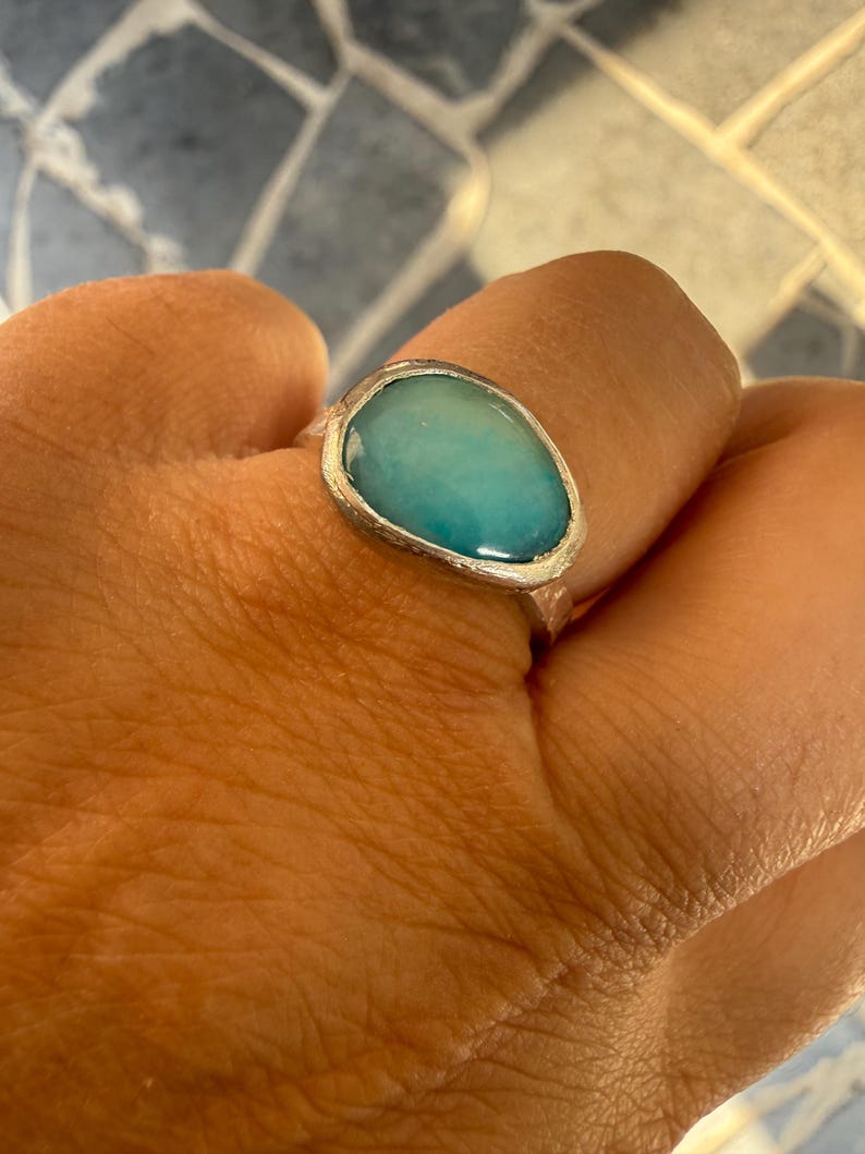 May include: A silver ring with a large, oval-shaped, light blue stone. The stone has a smooth, polished surface and is set in a textured silver band. The ring is displayed on a hand.