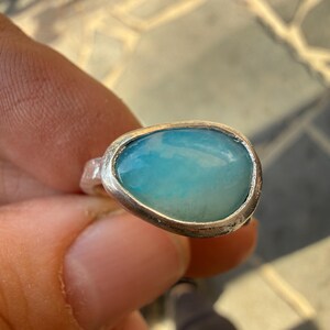 May include: A silver ring featuring a large, oval-shaped, light blue gemstone. The stone has a smooth, polished surface with subtle variations in color. The ring's band is a simple, unadorned design, highlighting the gemstone's natural beauty.