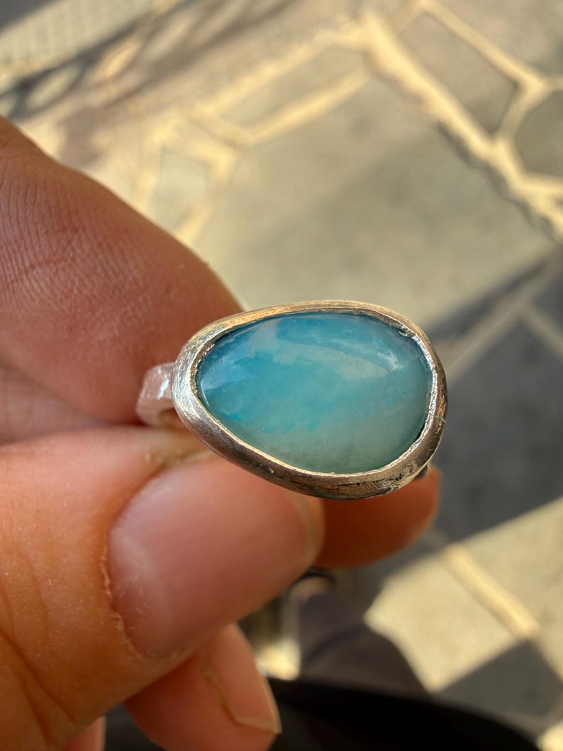 May include: A silver ring featuring a large, oval-shaped, light blue gemstone. The stone has a smooth, polished surface with subtle variations in color. The ring's band is a simple, unadorned design, highlighting the gemstone's natural beauty.