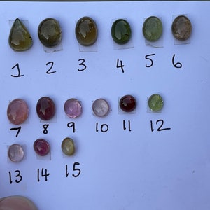 May include: A collection of fifteen loose gemstones in various shapes and colors, including shades of green, pink, red, and brown. Each stone is numbered from 1 to 15, arranged in three rows on a white surface, showcasing polished surfaces.