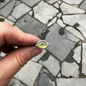 May include: A silver ring featuring a single, oval-shaped, faceted light green gemstone set in a smooth metal bezel. The band of the ring has a textured, slightly irregular finish, providing a handcrafted appearance to the piece of jewelry.