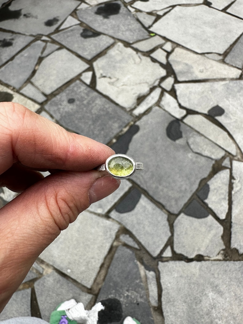 May include: A silver ring featuring a single, oval-shaped, faceted light green gemstone set in a smooth metal bezel. The band of the ring has a textured, slightly irregular finish, providing a handcrafted appearance to the piece of jewelry.