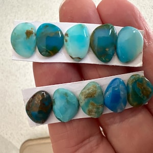 May include: A hand holds two strips of paper displaying turquoise gemstones. The stones vary in shades of blue and green, with some showing brown matrix patterns. The polished stones are oval and teardrop-shaped, ideal for jewelry making.