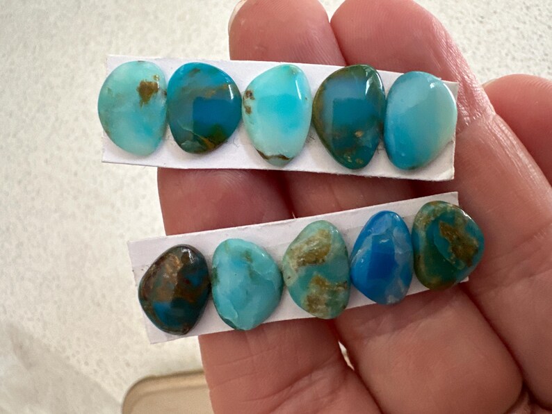 May include: A hand holds two strips of paper displaying turquoise gemstones. The stones vary in shades of blue and green, with some showing brown matrix patterns. The polished stones are oval and teardrop-shaped, ideal for jewelry making.