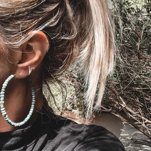 May include: A woman wearing a pair of large turquoise hoop earrings. The earrings are made of small, round beads and are about 7.5 cm in diameter.
