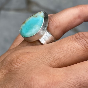 May include: A silver ring featuring a large, teardrop-shaped gemstone. The gemstone is a vibrant turquoise color with hints of brown and is set in a textured silver band. The ring is a statement piece, showcasing a unique and eye-catching design.