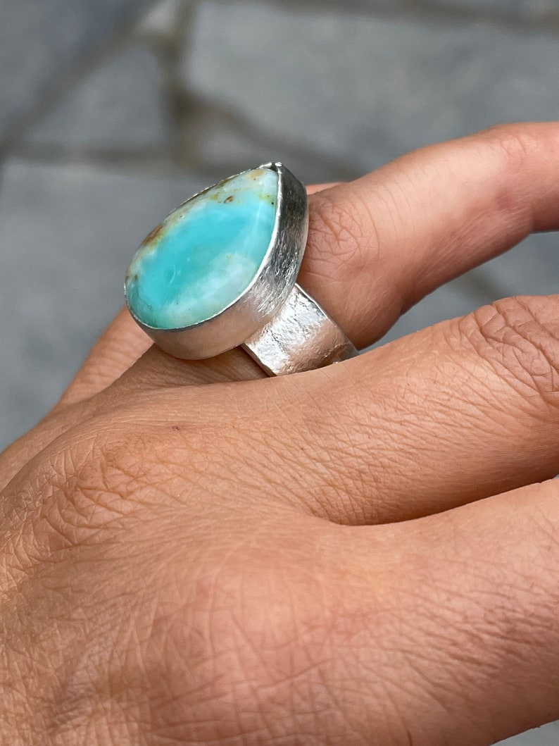 May include: A silver ring featuring a large, teardrop-shaped gemstone. The gemstone is a vibrant turquoise color with hints of brown and is set in a textured silver band. The ring is a statement piece, showcasing a unique and eye-catching design.