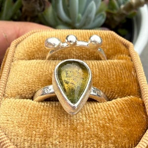 May include: Two silver rings resting in a mustard yellow velvet box. One ring features a teardrop-shaped olive green stone in a smooth bezel setting. The second ring has a textured band topped with three small, rounded silver spheres.