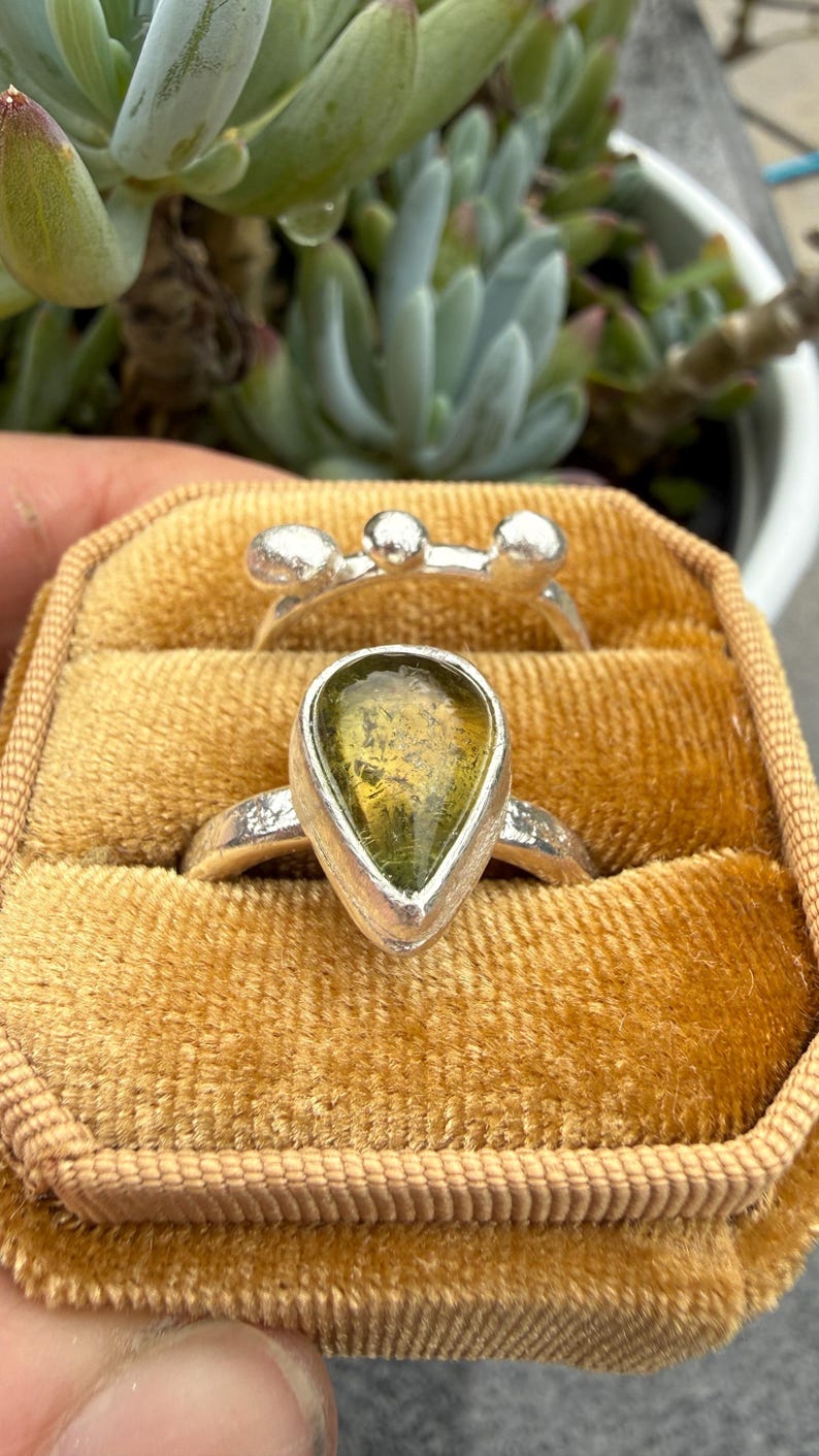 May include: Two silver rings resting in a mustard yellow velvet box. One ring features a teardrop-shaped olive green stone in a smooth bezel setting. The second ring has a textured band topped with three small, rounded silver spheres.