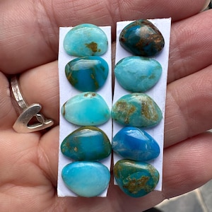 May include: A collection of turquoise gemstones in various shades of blue and green, with brown matrix patterns. The stones are oval-shaped and mounted on white paper strips. The image shows a close-up view of the gemstones.