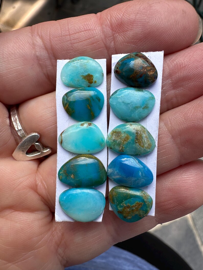 May include: A collection of turquoise gemstones in various shades of blue and green, with brown matrix patterns. The stones are oval-shaped and mounted on white paper strips. The image shows a close-up view of the gemstones.