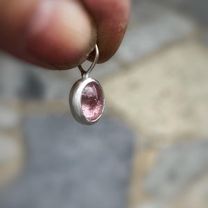 May include: A small oval pendant featuring a translucent pink gemstone set in a smooth, polished silver bezel. The pendant includes a simple silver jump ring at the top for attachment, showcasing a minimalist design with a focus on the stone.