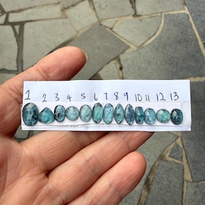 May include: Thirteen faceted blue-green gemstones with dark inclusions arranged in a row on a white paper strip labelled with numbers one through thirteen. Each stone displays unique shapes including oval, round, pear, and marquise cuts.