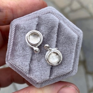 May include: Silver stud earrings with a circular design, displayed in a grey velvet jewellery box. The earrings feature a small, raised, round centre and a decorative drop.