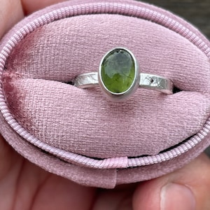 May include: A silver ring featuring an oval-shaped, translucent olive-green gemstone set in a smooth bezel. The ring band has a textured, hammered finish and is presented inside a soft, oval-shaped, light pink velvet ring box with a central slot.