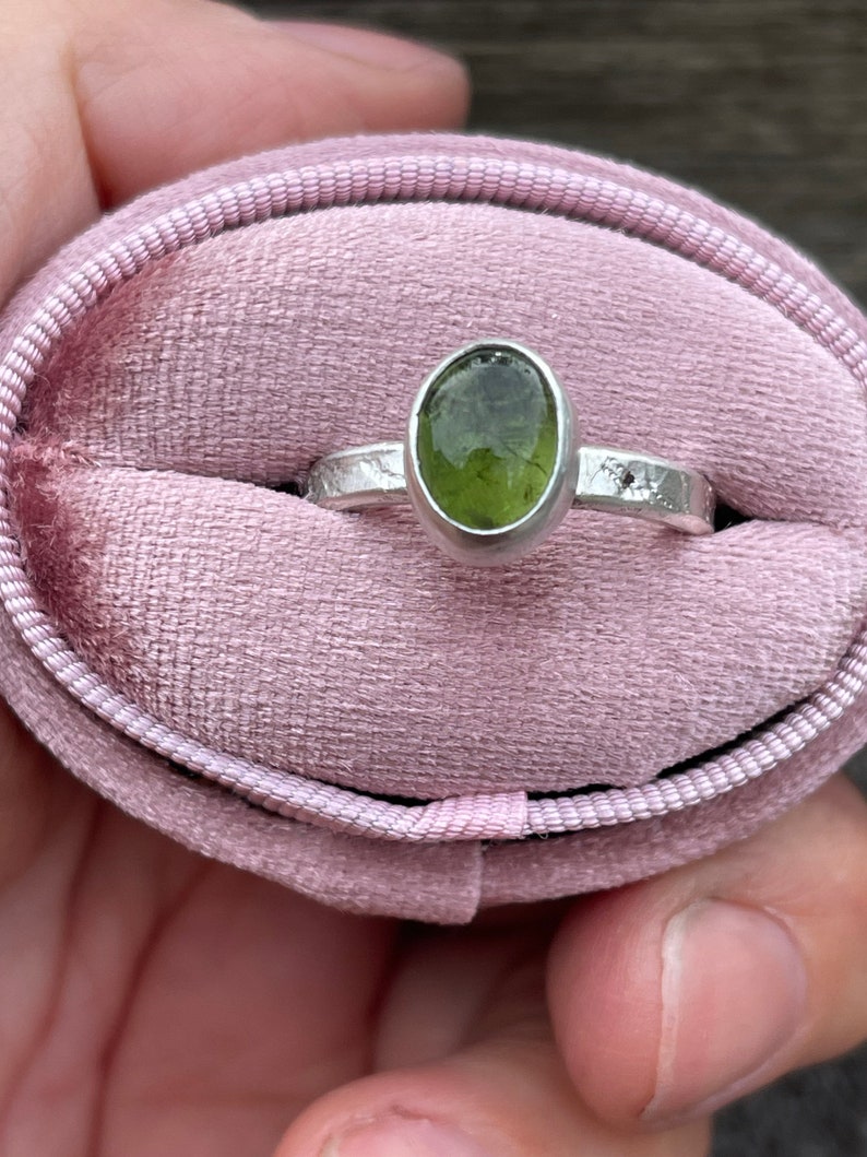 May include: A silver ring featuring an oval-shaped, translucent olive-green gemstone set in a smooth bezel. The ring band has a textured, hammered finish and is presented inside a soft, oval-shaped, light pink velvet ring box with a central slot.
