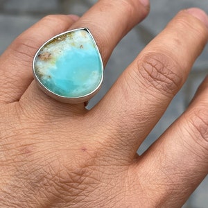 May include: A silver ring featuring a large, teardrop-shaped gemstone. The stone has a light blue center with brown and white inclusions along the top edge. The ring is set in a polished silver band.