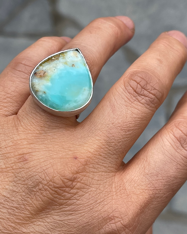 May include: A silver ring featuring a large, teardrop-shaped gemstone. The stone has a light blue center with brown and white inclusions along the top edge. The ring is set in a polished silver band.