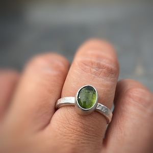 May include: A silver ring featuring an oval-shaped, translucent olive-green gemstone set in a smooth bezel. The metal band has a textured, hammered finish that wraps around the finger, providing a distinct contrast to the polished green stone.