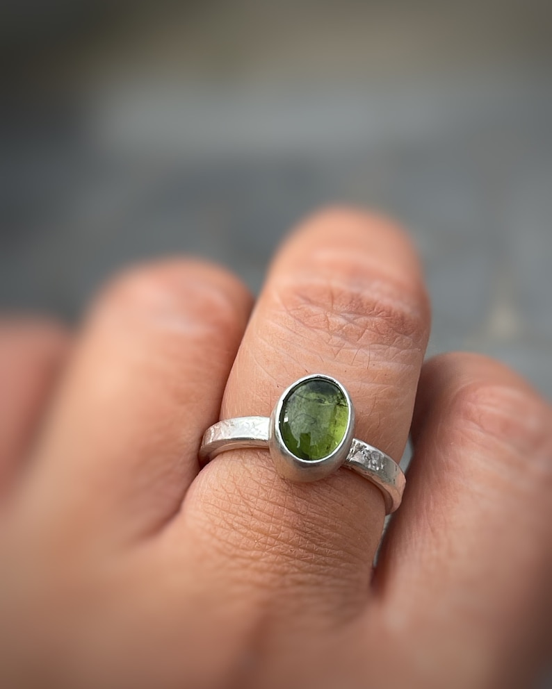 May include: A silver ring featuring an oval-shaped, translucent olive-green gemstone set in a smooth bezel. The metal band has a textured, hammered finish that wraps around the finger, providing a distinct contrast to the polished green stone.