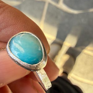 May include: A silver ring featuring a large, oval-shaped, light blue gemstone. The gemstone has a smooth, polished surface and is set in a textured silver bezel. The ring's band is also textured.