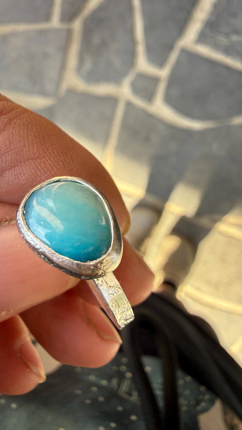 May include: A silver ring featuring a large, oval-shaped, light blue gemstone. The gemstone has a smooth, polished surface and is set in a textured silver bezel. The ring's band is also textured.
