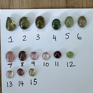 May include: A collection of fifteen polished, cabochon-cut gemstones arranged in three rows on a white sheet of paper. Each stone is numbered from 1 to 15, displaying various colors including shades of green, brown, pink, and deep red.
