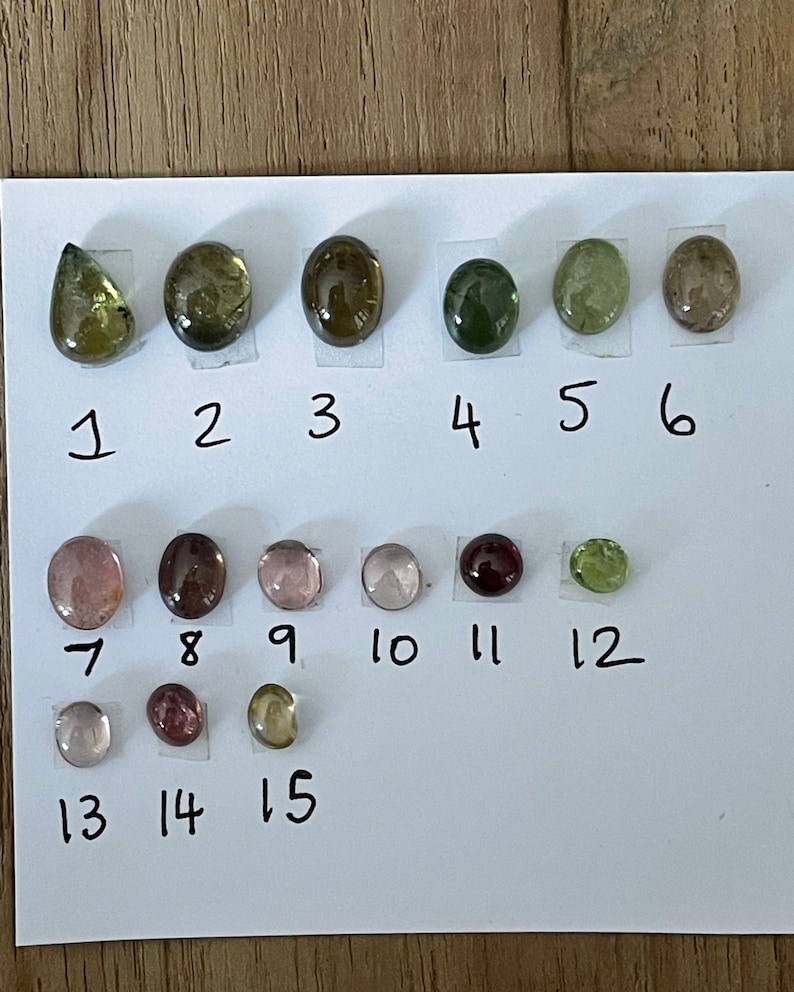 May include: A collection of fifteen polished, cabochon-cut gemstones arranged in three rows on a white sheet of paper. Each stone is numbered from 1 to 15, displaying various colors including shades of green, brown, pink, and deep red.