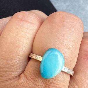 May include: A silver ring with a large, oval, light blue gemstone. The band has a hammered texture. The ring is worn on a finger, with the skin visible in the background. The gemstone is smooth and polished.