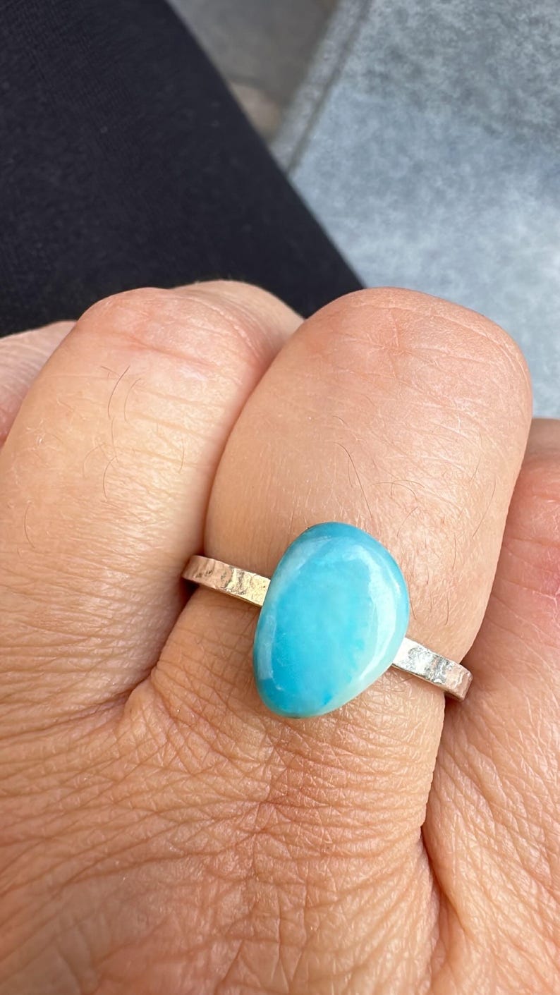 May include: A silver ring with a large, oval, light blue gemstone. The band has a hammered texture. The ring is worn on a finger, with the skin visible in the background. The gemstone is smooth and polished.