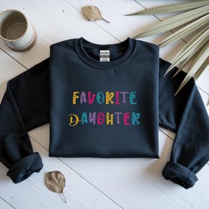 Op de afbeelding: Een marineblauwe sweatshirt met de tekst "FAVORITE DAUGHTER" in kleurrijke, geborduurde letters. De sweatshirt wordt gepresenteerd op een wit houten oppervlak, met decoratieve bladeren en een keramische mok op de achtergrond.